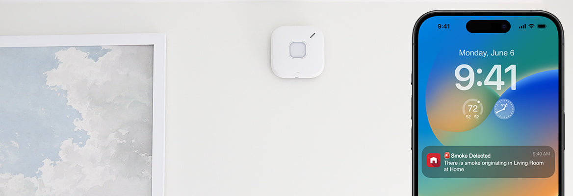 SC5 Smart Smoke & Carbon Monoxide Alarm on a wall near a painting of clouds. A smartphone shows an app notification of smoke detected in the living room.